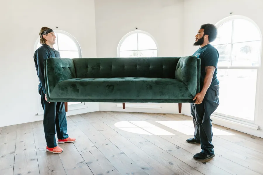 Two movers carefully carrying a green velvet sofa into a bright room